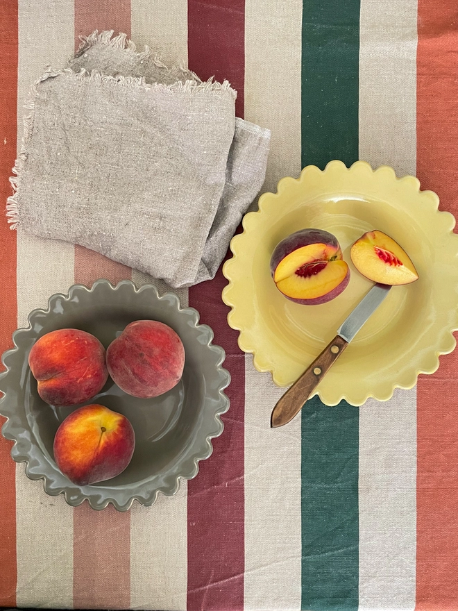 peaches shown on a grey and a yellow shallow bowl with scalloped edge on a striped cloth