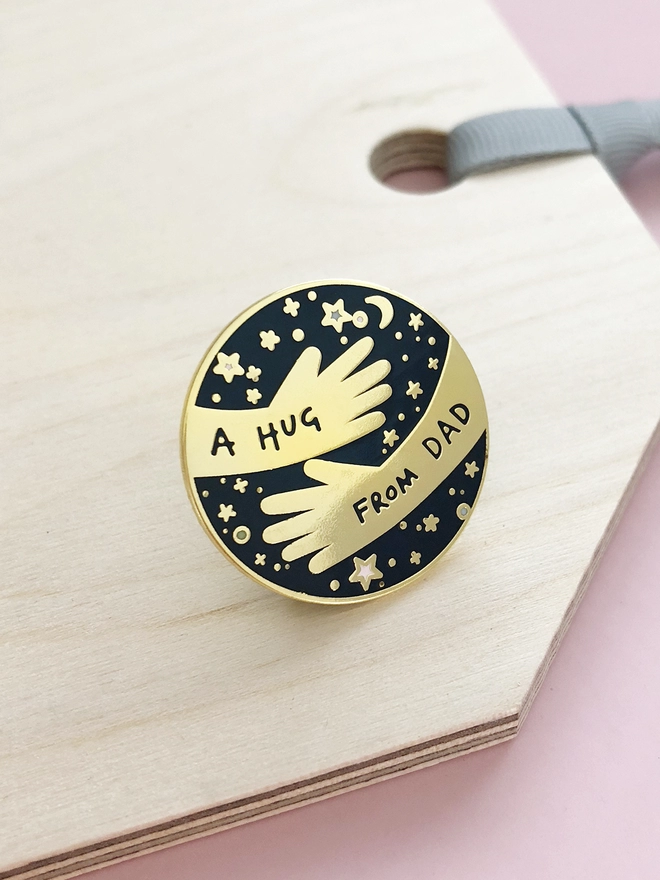 A navy blue and gold pin badge with a hugging arms design and the words “A hug from Dad” is placed on a wooden tag.