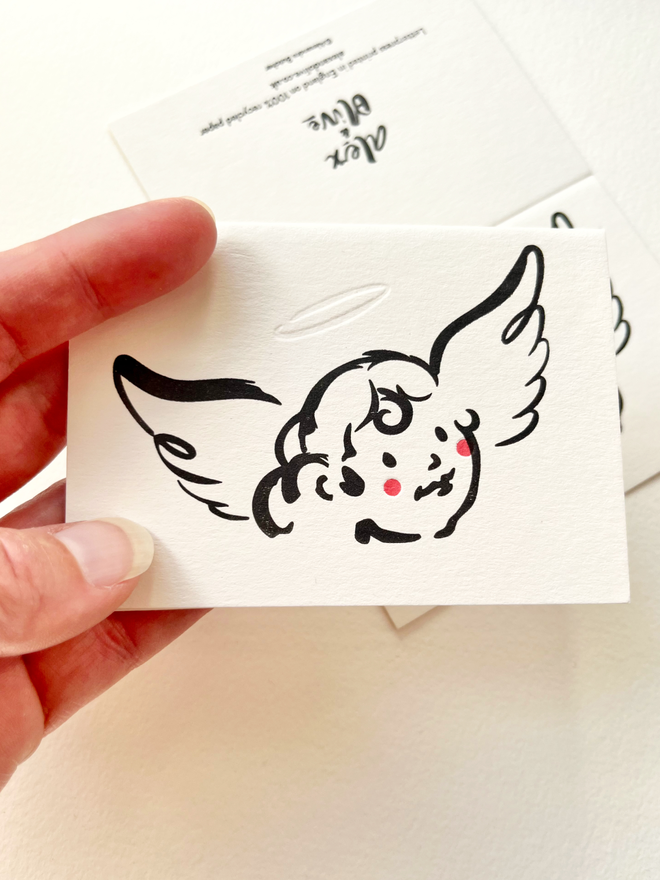 small greeting card featuring a simple christmas cherub character in black ink being held in the hand. 