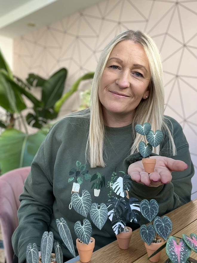 Nicole holding a miniature replica Philodendron Gloriosum paper plant ornament in a terracotta pot on the palm of her hand with other paper plants in the foreground