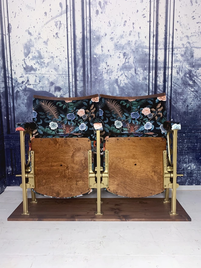 A set of two vintage cinema seats upholstered in a black floral velvet against a blue marbled wall.  Both seats are up