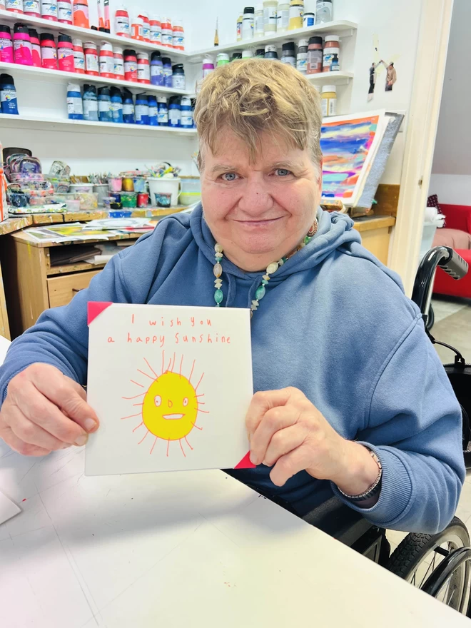 Artist holding I Wish You A Happy Sunshine card with a sketch of a smiling yellow sun outlined in orange