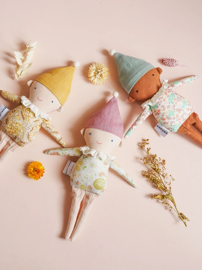 Three handmade Spring Elf Dolls are sitting side by side against a soft pastel background. Each doll has a floral print body, pastel-coloured hat with a pompom, and a ruffled white collar. They feature different skin tones and gentle smiles, radiating charm and warmth.