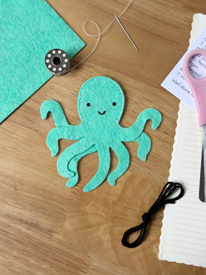 A turquoise felt octopus finger puppet is on a wooden desk beside the craft kit materials that have been used to make it.