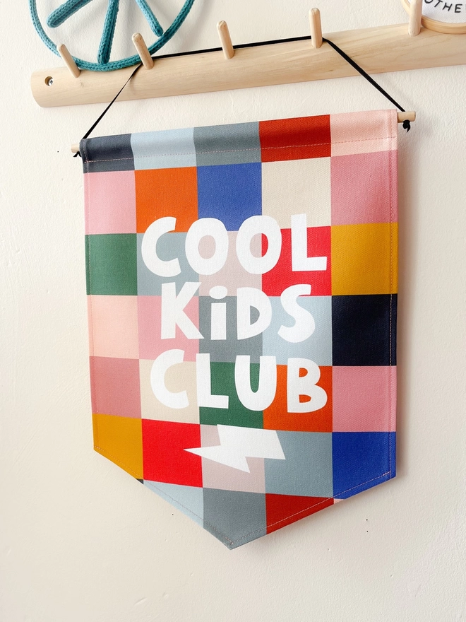 Wall banner hanging from a wooden peg rail saying the words cool kids club, in various coloured chequered background