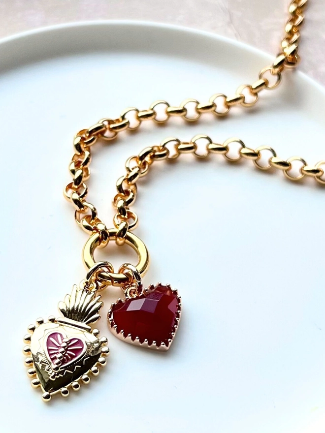 Gold and red enamel heart charm with red quartz heart charm hanging from gold belcher chain necklace on a white dish