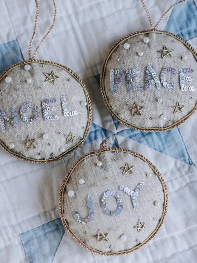embroidered linen peace joy and noel decorations. beige linen hanging decorations laid on blue and white quilt.
