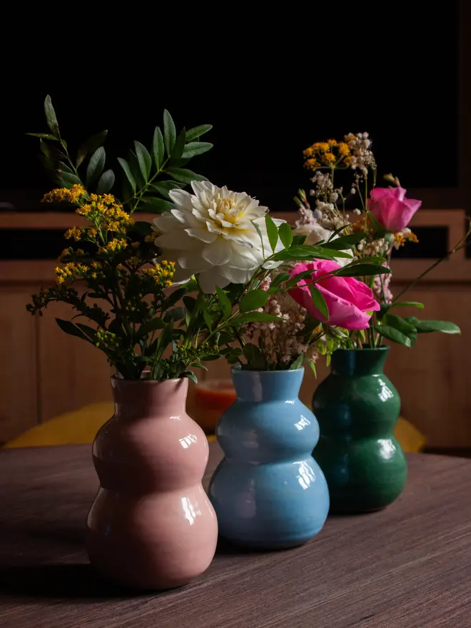 Chubby Bud Vases in a row of three colours sitting on a wooden table. the bud vases are holding a small bunch of flowers and are in pink, green and blue