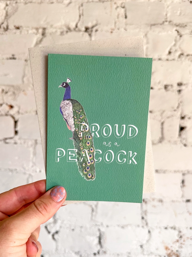 a deep green greetings card featuring an illustration of a peacock with the text “proud of a peacock”