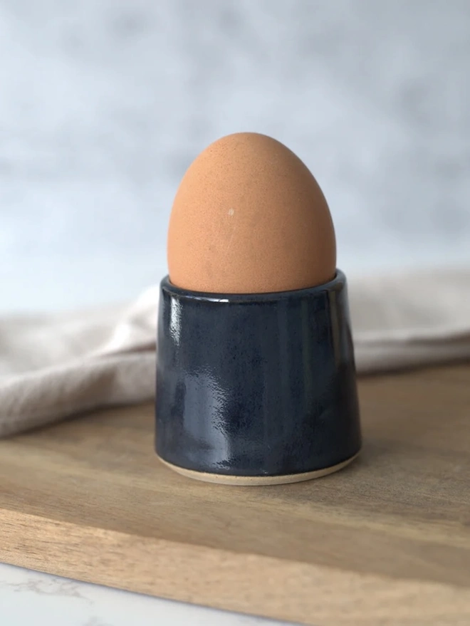 Classic Handmade Egg Cup, a ceramic egg cup sitting on a wooden chopping board against a grey backdrop. 