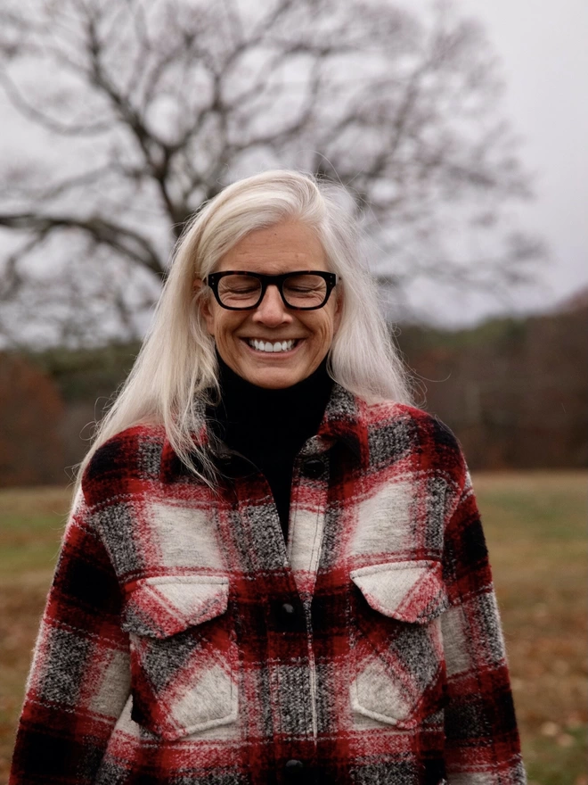 Woman with long grey hair wearing a checked jacket smiling with her eyes closed standing in a park