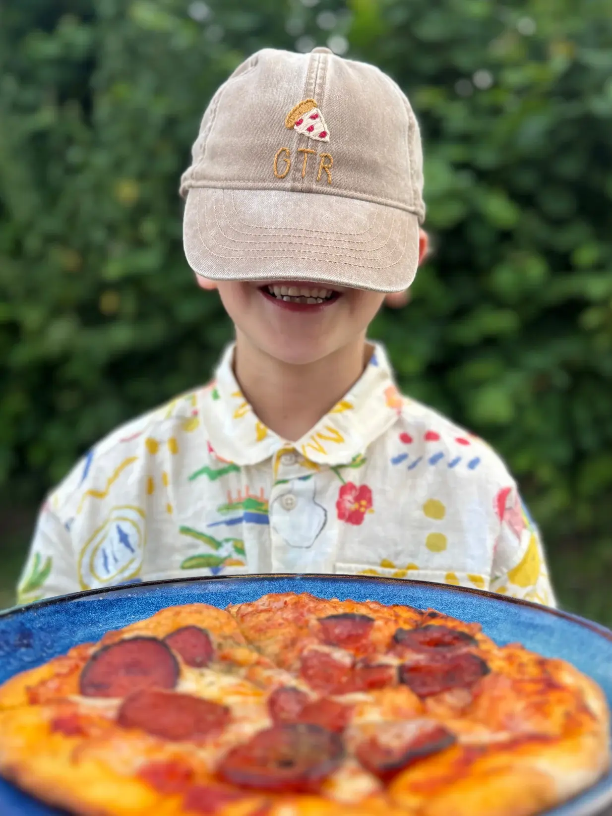Hand Embroidered Kids Cap, a child standing outside with a personalised cap. He is accompanied by a pizza. 