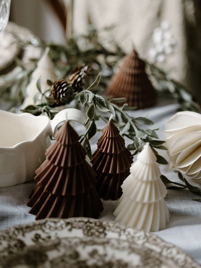 Brown Christmas Tree Warm Candles (Set Of 2). Brown and cream ribbed Christmas tree candles on a linen tablecloth, with eucalyptus, pine cones, a paper bauble and a vintage floral plate. The small sculptural candles make charming table favours or stocking fillers.