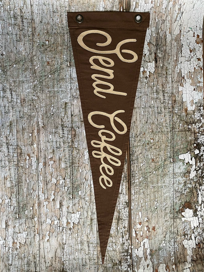 An espresso brown canvas pennant flag hung vertically on a wooden wall with the words 'Send Coffee' in a latte coloured canvas.