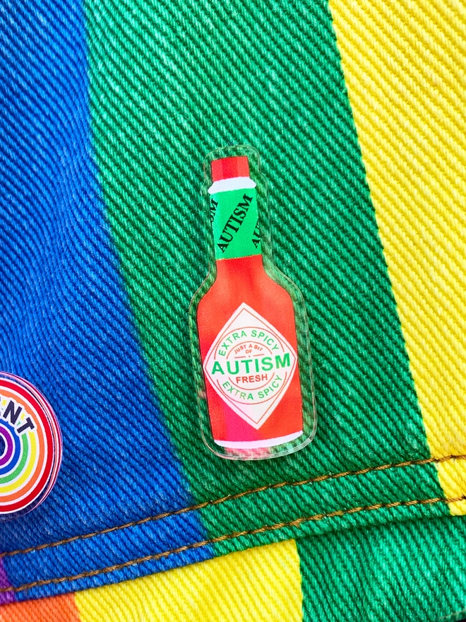 an acrylic badge shaped and designed to look like a miniature hot sauce bottle. the bottle says 'fresh autism' on the label. the badge is pinned to a rainbow striped denim jacket.