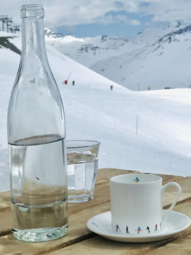 Ski Chain Espresso Cup. An espresso cup is seen on a wooden table in front of a snowy mountain scene. The white cup is patterned on its lower portion with a simple, multicoloured chain of skiers.
