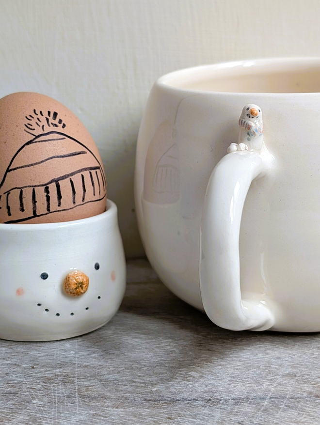 white cup with a tiny ceramic snowman and snowballs on the handle next to a pottery snowman egg cup with an egg in