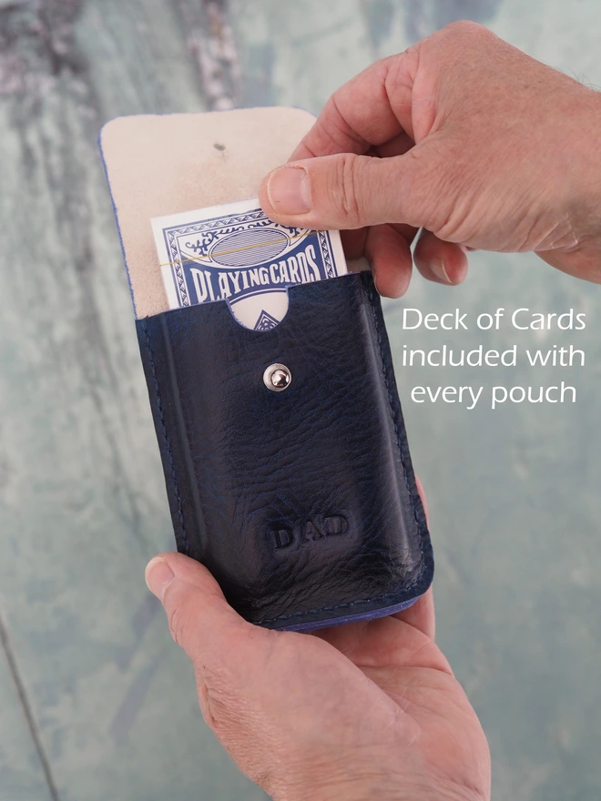 Personalised leather playing card holder. A dark blue handmade leather sleeve with a new deck of cards inside, being slid out by a pair of hands.