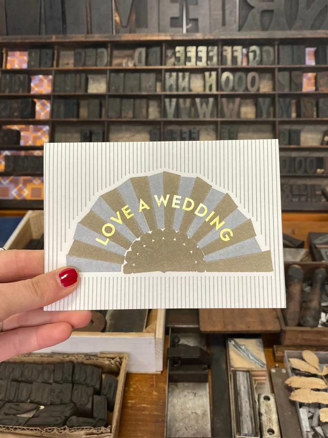 A persons hand holds a white and silver striped card with silver and gold fan on it and gold text reading 'Love A Wedding'.