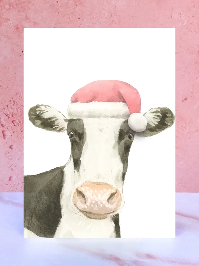 A Christmas card featuring a hand painted design of a cow, stood upright on a marble surface surrounded by pompoms. 