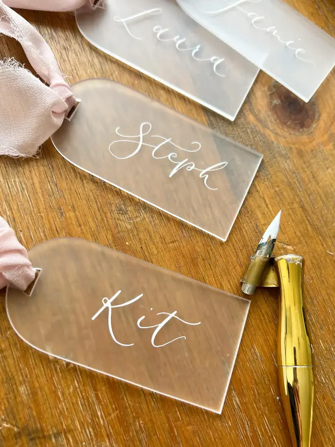 Personalised Calligraphy Frosted Acrylic Place Cards, frosted acrylic place cards with pink ribbons grouped together on a wooden table. 