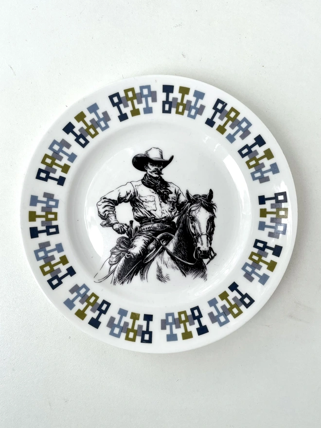 vintage plate hanging on a white wall, it has a decorative border of grey and blue leaves, with a printed vintage illustration of a cowboy in the middle 