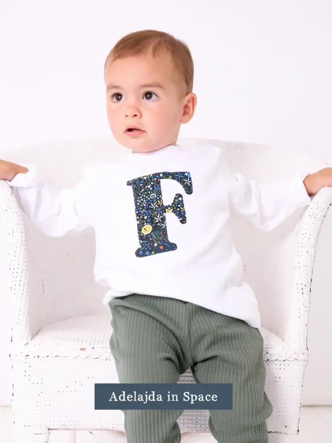 a 1 year old boy wearing a personalised kids white t-shirt with an initial cut from liberty space print on the front