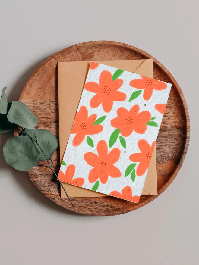 Seed Card with orange floral illustrations all over placed on a wooden tray next to a Eucalyptus branch