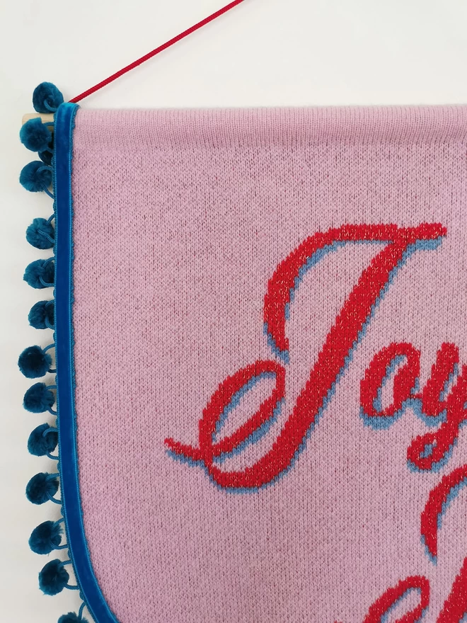 A close up product image of the front of a pink knitted scallop shaped wall hanging with teal pom pom trim. The start of the words ‘Joyeux Noel’ are written across the banner in red retro writing with a hint of gold sparkle in the text and a blue shadow to the lettering .You can see the letter ‘J’ here in detail.