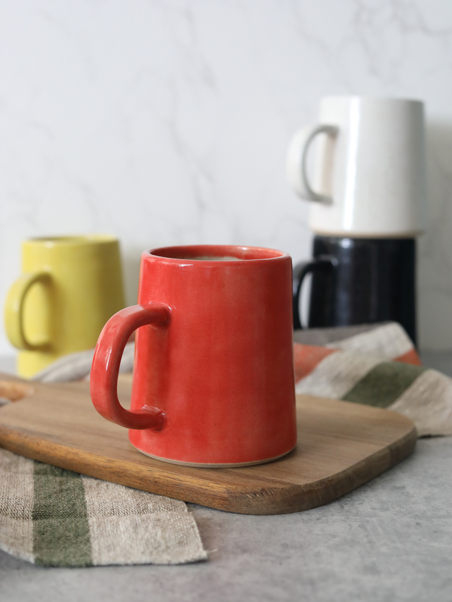 A cherry red handmade ceramic pint mug filled with a hot drink sits on a wooden board in the foreground, with yellow, black, and speckled white mugs softly blurred behind, styled with linen cloths against a white marble background