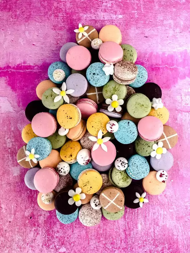 a pile of colourful easter macarons arranged in the shape of an easter egg on a pink background with daisy flowers and mini meringues