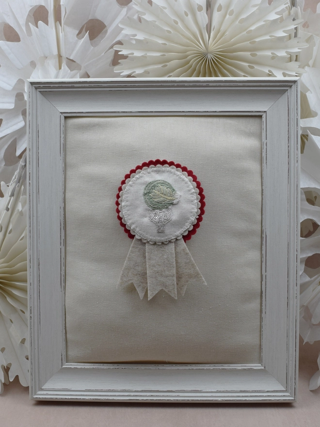 Christmas red and Beige felt rosette with embroidered Brussels Sprout on a fork as the central design.  Pinned to a padded frame with paper snowflakes in the background.