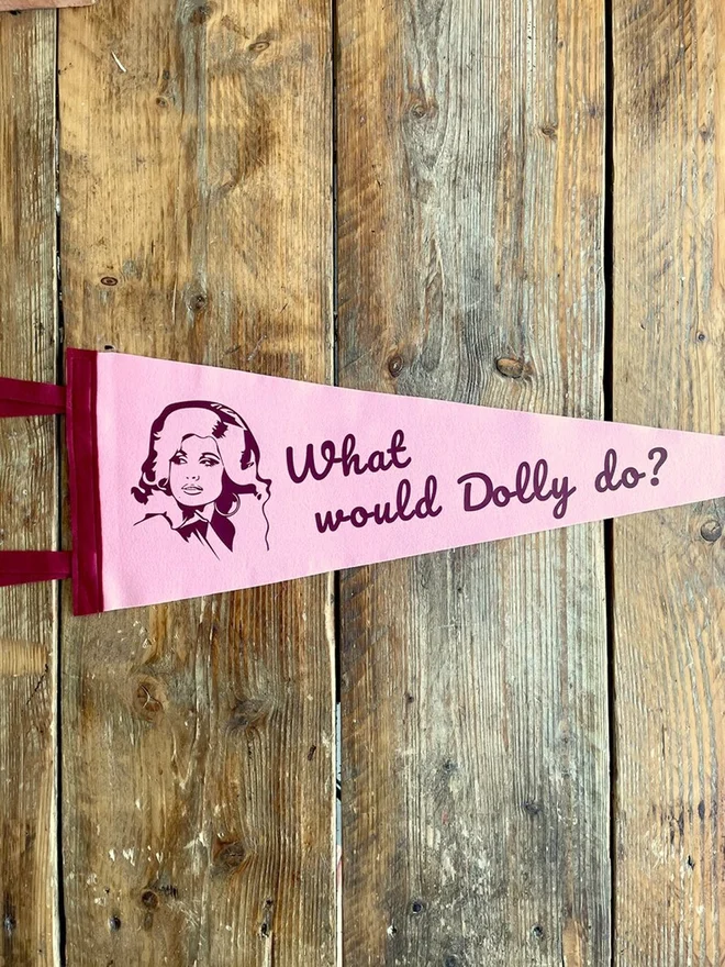 A pastel pink felt pennant with burgundy trim and ties, printed in burgundy the beautiful face of country adn western singer Dolly Parton and the phrase what would Dolly do