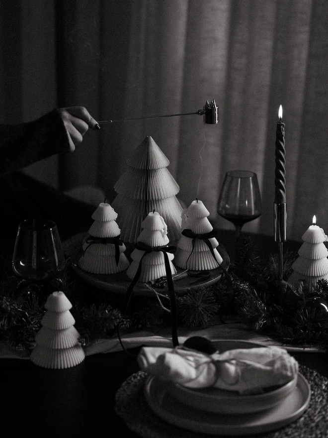 Tiered Christmas Tree Candle. A black and white image of a hand using a candle snuffer over cream tree candles with velvet bows, on a moody Christmas dinner table with a twisted taper candle and wine glasses. The timeless scene shows how elegantly the sculptural candles suit festive tablescapes.
