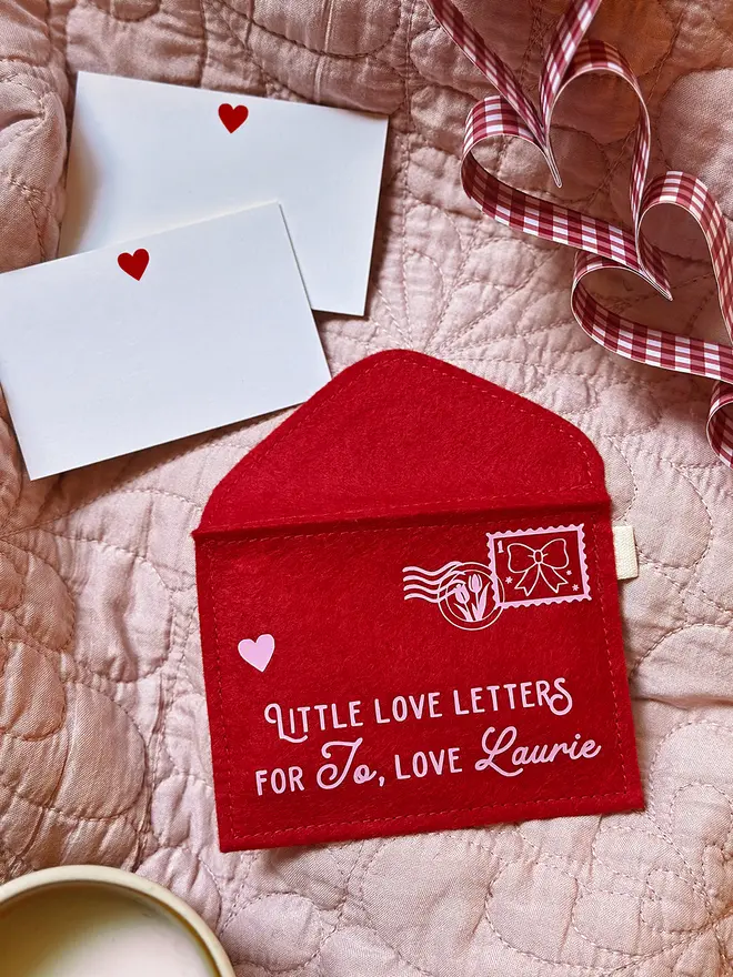 A personalised red felt Valentine’s envelope with pale pink lettering reading “Little Love Letters”, shown with two small heart cards on a soft pink quilt and gingham ribbon, styled as a romantic handmade keepsake.