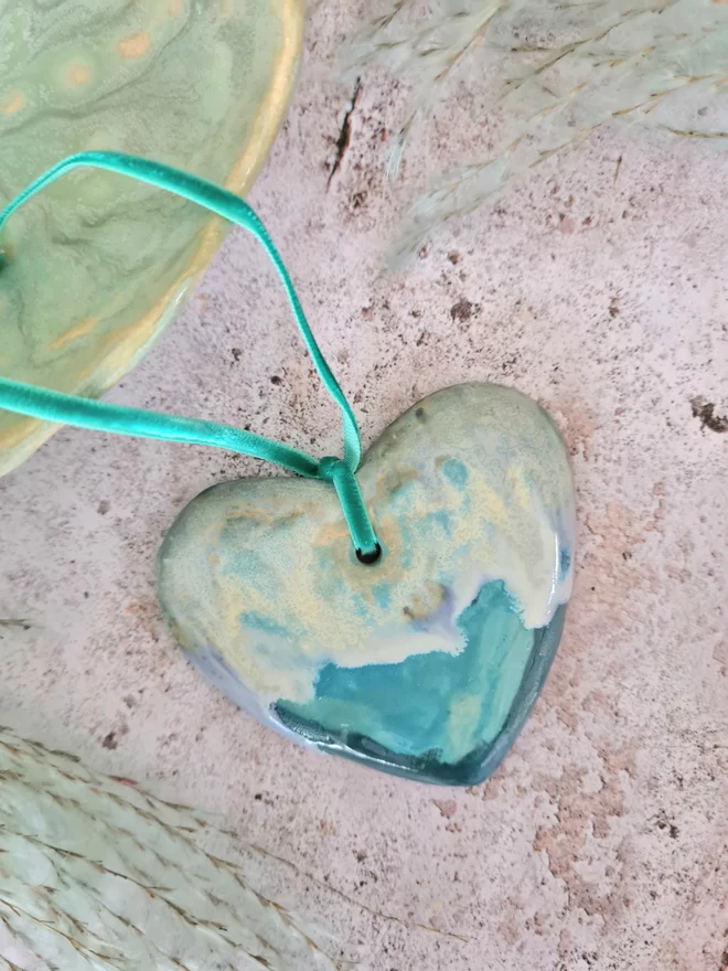 ceramic hanging heart decoration with blue, green, turquoise, Jenny Hopps Pottery, velvet ribbon, gift, mothers day gift, valentines gift, homeware, photographed hanging from a grey backdrop with dried flowers.