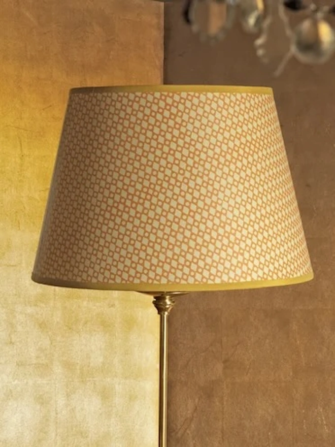 Retro Drum Lampshade. A tapered drum lampshade in a peach and cream diamond lattice block print with a mustard yellow trim, fitted to a brass floor lamp. The softly patterned shade glows warmly against a gilded wall for an elegant living room setting.