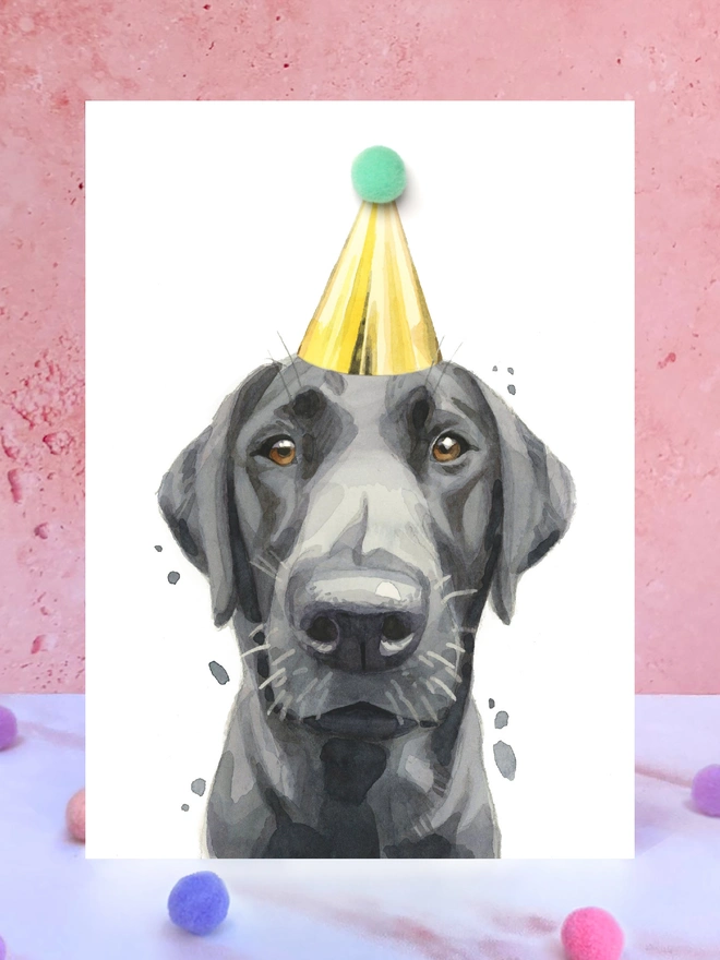 A greeting card featuring a hand painted design of a black labrador, stood upright on a marble surface surrounded by pompoms. 
