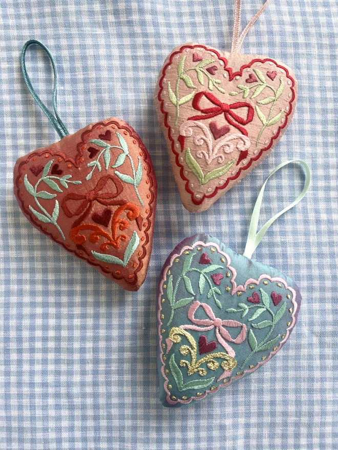 three silk ornaments with hand-guided embroidery including bows, leaves, hearts and a scalloped edge