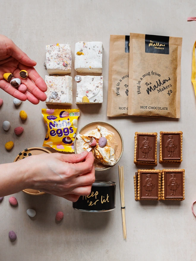 looking down on a hand putting crushed mini eggs on top of hot chocolate with contents of The Mallow Makers Easter luxury s'mores and hot chocolate kit
