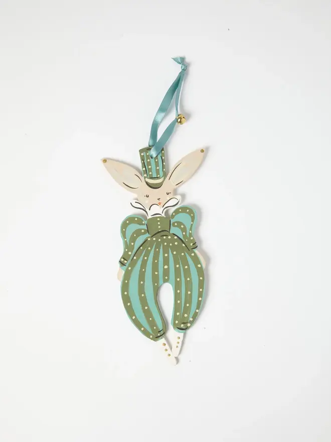 Verdant Hare Tree Dancer. a flat lay product shot of the verdant hare tree dancer hanging ornament on a plain white background, showing a hare figure in a striped green and teal jumpsuit with a matching top hat and bow tie. the figure hangs from a teal ribbon with a small gold bell attached, its ears standing tall above a serene, closed-eye expression. hand-painted polka dot detailing gives the ornament a folk-art, storybook charm.