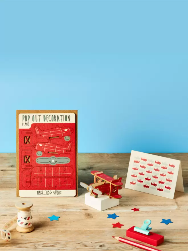 3D laser-cut red biplane decoration and plane pattern greeting card and brown kraft envelope on top of a wooden desk in front of a blue coloured background