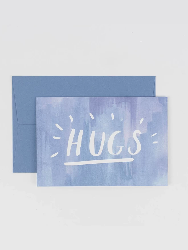 A hand lettered card featuring the word "HUGS" in capital letters. On a pale blue watercolour background