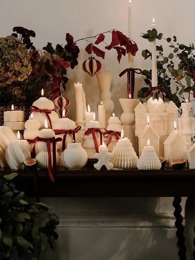 White Gingerbread Candles (Set Of 2). A console table covered in lit cream sculptural candles, including houses, trees, a gingerbread man, a nutcracker and ribbed pillars tied with red velvet bows, styled with dried hydrangeas and foliage. The glowing collection creates a magical, festive centrepiece for the holiday season.