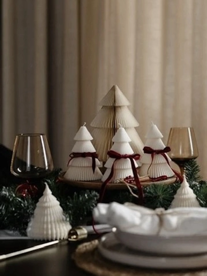 Tiered Christmas Tree Candle. Three lit cream Christmas tree candles with ribbed tiers and burgundy velvet bows, arranged on a wooden board in front of a paper honeycomb tree. Pine garland, cinnamon sticks and red berries complete this cosy festive centrepiece.