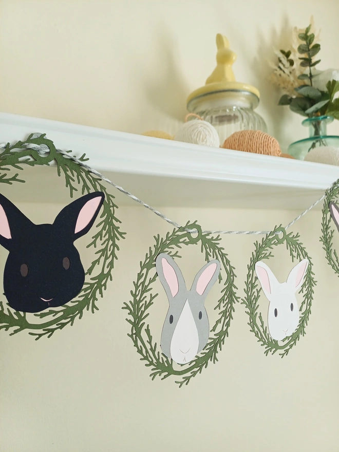 Easter Bunny Paper Garland closeup showing Black bunny decoration, light grey and white bunny, and white bunny decoration. All rabbits have pale pink ears and are surrounded by olive green foliage