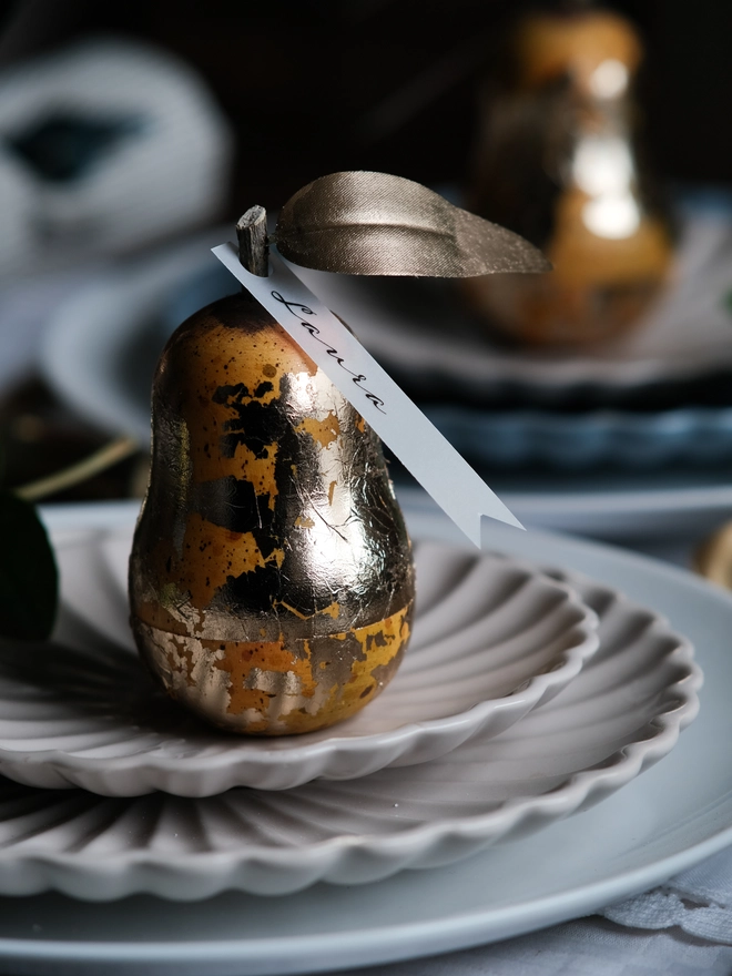 Gilded vintage-style Christmas pear ornament opened to reveal a small surprise inside, shown on a festive Christmas table setting with white plates and ribbon.