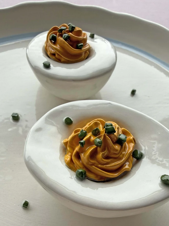 Ceramic devilled eggs with chive detail, on a plain white plate with blue trim, on a pink background