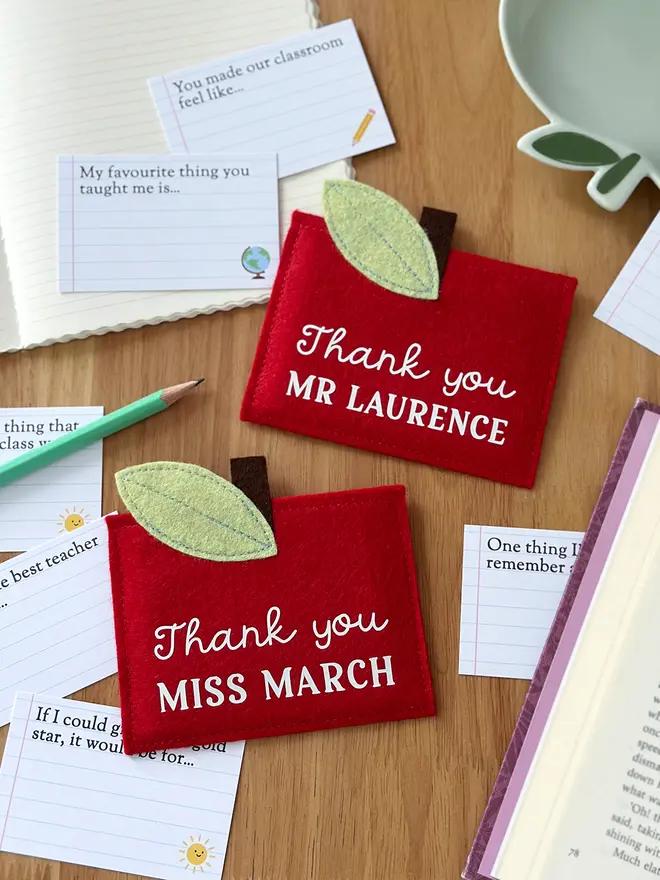 Two handmade red felt apple-shaped envelopes with green felt leaves and brown stems, each reading "Thank you MR LAURENCE" and "Thank you MISS MARCH", surrounded by illustrated prompt cards with child-friendly questions, displayed on a wooden desk with a pencil, notebook, and book. It's a charming personalised, handmade teacher thank you gift.