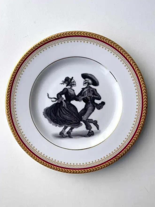 vintage plate with an ornate border, with a printed vintage illustration of two dancing day of the dead skeletons wearing a dress and a suit in the middle